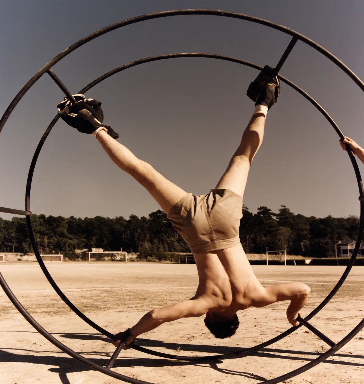 A gymnast performing inside a large metal wheel on a field, with trees along the horizon.