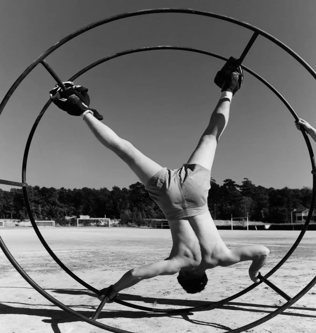 A grayscale photograph of a gymnast performing inside a large metal wheel on a field.